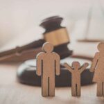 Ícones de madeira representando uma família, com duas figuras adultas e uma figura infantil ao centro, posicionados sobre uma superfície de madeira. Ao fundo, desfocados, aparecem um martelo de juiz e uma balança da justiça, remetendo a temas jurídicos, direito de família ou processos judiciais. A imagem possui iluminação suave em tons quentes e profundidade de campo reduzida.