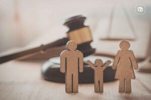 Ícones de madeira representando uma família, com duas figuras adultas e uma figura infantil ao centro, posicionados sobre uma superfície de madeira. Ao fundo, desfocados, aparecem um martelo de juiz e uma balança da justiça, remetendo a temas jurídicos, direito de família ou processos judiciais. A imagem possui iluminação suave em tons quentes e profundidade de campo reduzida.